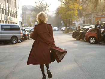 Young woman with curly hair walking on city street at sunset wearing red coat