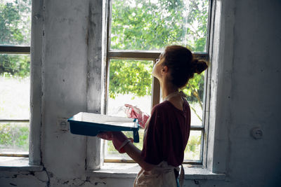 Side view of woman holding book while standing against window