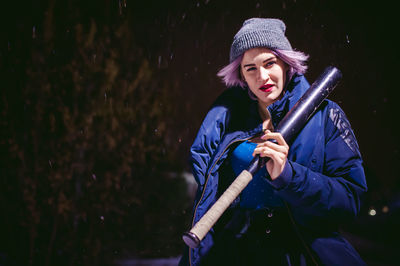 Young woman with baseball bat standing outdoors at night
