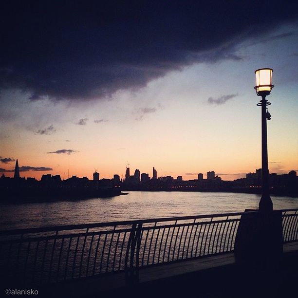 architecture, built structure, building exterior, silhouette, city, sky, sunset, water, river, railing, cityscape, street light, sea, tower, illuminated, skyscraper, dusk, lighting equipment, cloud - sky, communications tower