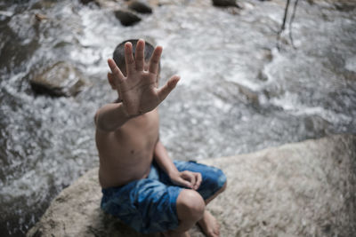 Full length of shirtless boy covering face with hand while sitting on rock in river