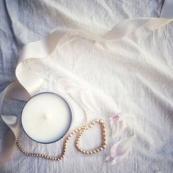 Directly above view of scented candle and pearl jewelry with ribbon on white bed