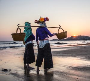 Rear view of friends with baskets standing at beach during sunset