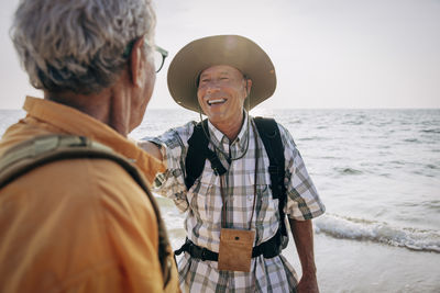Happy senior gay man talking with boyfriend while standing near sea at beach on vacation