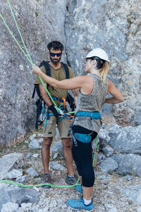 Fearless active couple in safety harnesses preparing gear for ascending rocky mountain slope while practicing climbing in nature