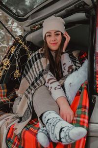 Brunette woman wrapped in blanket in trunk car drinking coffee tea from thermos. travel in winter. 