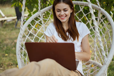 Portrait of young woman using laptop while standing by chainlink fence