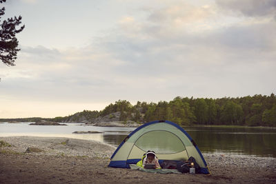 Girl using digital tablet while lying in tent at beach against sky during sunset