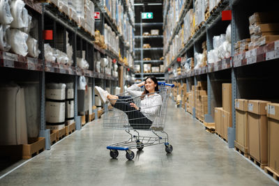 Full length of young woman sitting on shopping cart in warehouse