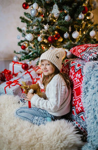 Happy little girl in white hat holding christmas gift boxes while sitting on floor.