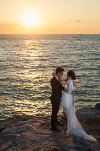 Couple standing on rocks by sea during sunset