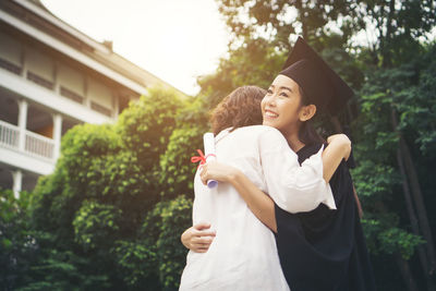 Cheerful woman in graduation gown embracing mother while standing outdoors