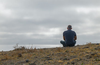 Rear view of adult man sitting on hill against cloudy sky and sea. cabo de gata park, almeria, spain