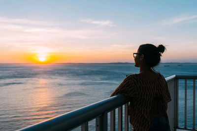 Woman standing by railing against sea during sunset