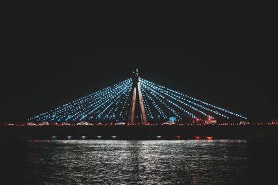 Illuminated suspension bridge over river against sky at night