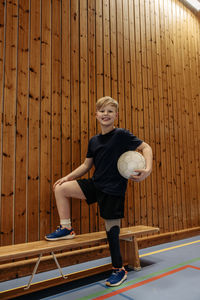 Smiling boy with amputated leg holding soccer ball while standing by wooden wall at sports court