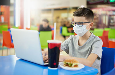 Teenager wearing glasses and a protective mask uses a laptop in a restaurant before lunch.