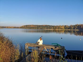 Rear view of woman with dog relaxing on lake pier against clear sky