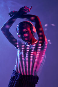 Charming brazilian female listening to music in wireless headphones while dancing on purple background in studio with shadows on disco ball and fluorescent lights