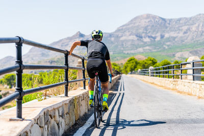 Back view of unrecognizable bicyclist in sportswear and helmet resting near railing while riding bike up paved road in rural mountainous area