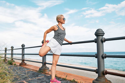 Young beautiful sportive woman training over seaside promenade, stretching legs before jogging.