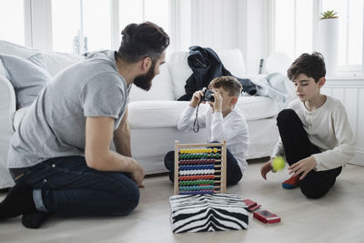 Boy photographing father by brother playing with ball in living room at home