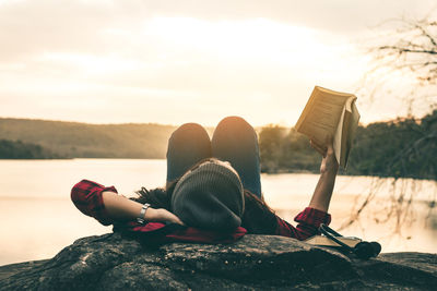 Woman reading book while lying on lakeshore during sunset