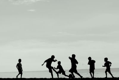 Silhouette boys playing soccer at beach against sky