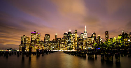 Illuminated modern buildings by river against sky at night