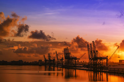 Silhouette cranes at harbor against sky during sunset