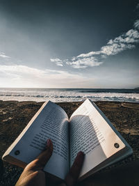 Midsection of person reading book on beach against sky