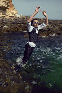 Man with a beard and sunglasses in clothes a vest and a white shirt jump in the sea among the rocks