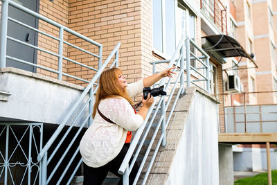 Smiling young woman holding camera standing against steps