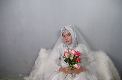 Portrait of bride wearing traditional clothing holding bouquet against wall
