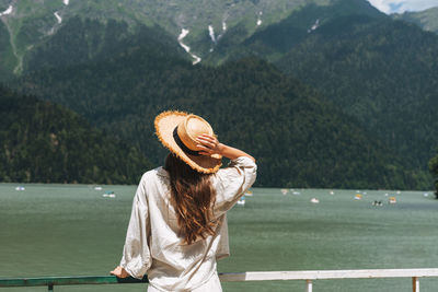 Young woman traveler with long blonde hair in straw hat looks at beautiful view of mountain lake