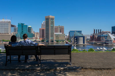 Rear view of couple sitting on bench in city against clear sky