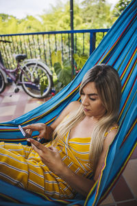 Serious young female in yellow sundress using smartphone and chilling in hammock on resort terrace