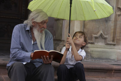 Full length of grandfather reading story to boy with umbrella outdoors