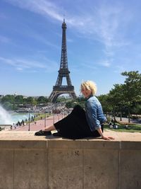 Woman sitting on railing with eiffel tower in background against blue sky