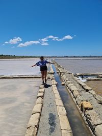 Woman with arms outstretched walking on rock by lake against blue sky