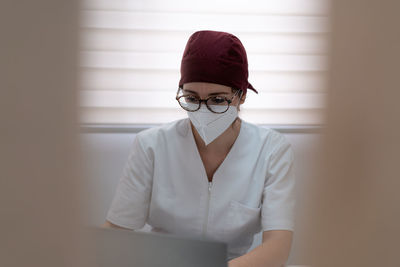 Concentrated female doctor in medical mask and uniform sitting at table and browsing laptop while working in modern workspace