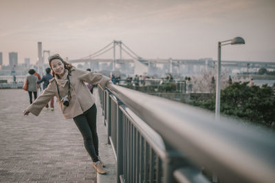 Portrait of happy woman holding railing while standing against bridge in city during sunset