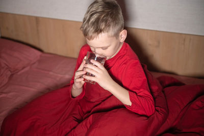 A happy boy is sitting in red pajamas under a blanket and drinking water from a glass