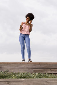 Low angle view of smiling woman listening music on mobile phone while standing on wood against sky