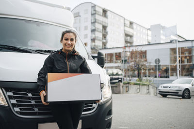 Portrait of smiling female worker carrying box while standing against delivery van in city