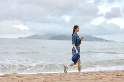 Side view of young woman standing at beach against sky