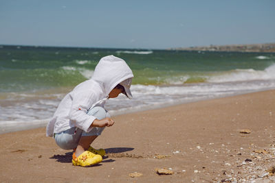 Baby boy sit on the beach with sand in yellow slippers and white shirt