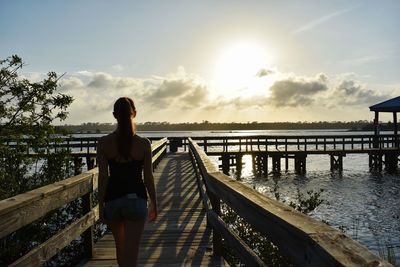 Rear view of woman standing on footbridge against sky during sunset