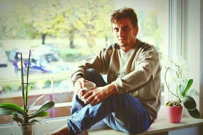 Portrait of young man holding coffee cup while sitting on window sill at home