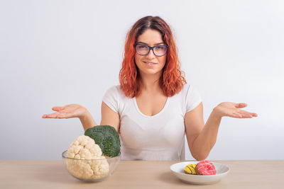 Redhead girl chooses between broccoli and donuts on white background.